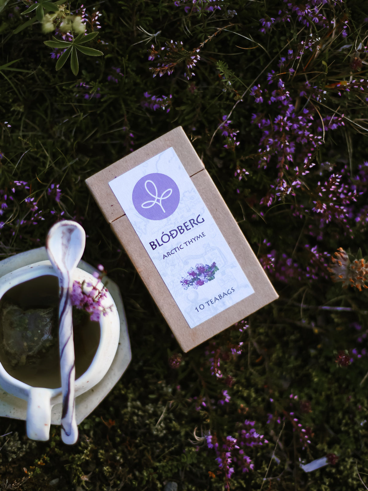 A kraft paper tea box labeled 'BLOBBERG Arctic Thyme' lies on a bed of green moss and small purple flowers, with a white ceramic teacup and spoon nearby.