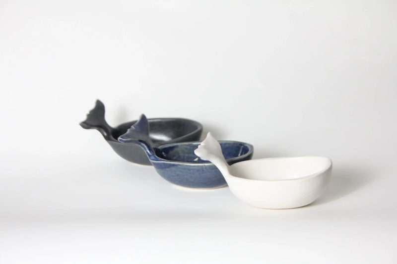 Three ceramic whale-shaped bowls in dark gray, medium blue, and white, arranged diagonally on a seamless light gray background.